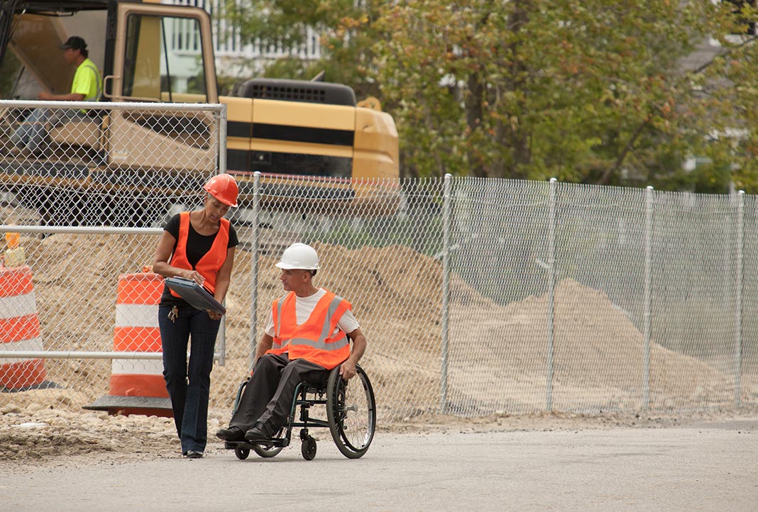 Construction woman showing clipboard to construction man in a wheelchair at a jobsite